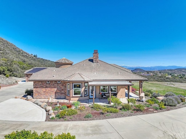 back of property with a tile roof, stone siding, a patio, a mountain view, and a chimney