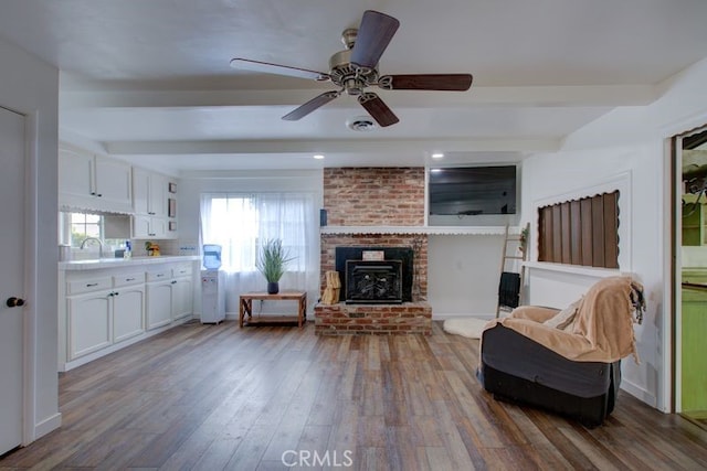 living area featuring a ceiling fan, beam ceiling, a brick fireplace, and wood finished floors