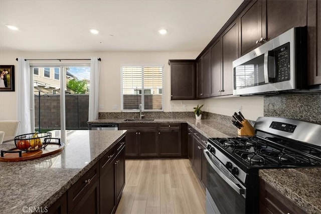 kitchen with dark stone countertops, a sink, dark brown cabinetry, appliances with stainless steel finishes, and light wood-type flooring