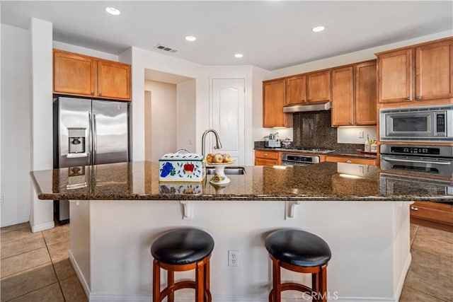 kitchen featuring under cabinet range hood, a sink, stainless steel appliances, a breakfast bar area, and brown cabinetry