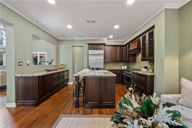 kitchen featuring dark brown cabinets, glass insert cabinets, premium range hood, built in appliances, and a sink