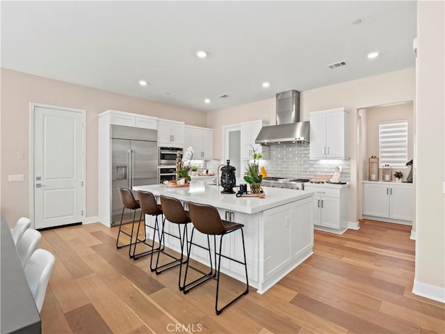 kitchen featuring visible vents, stainless steel built in refrigerator, a breakfast bar area, light wood-style flooring, and wall chimney exhaust hood