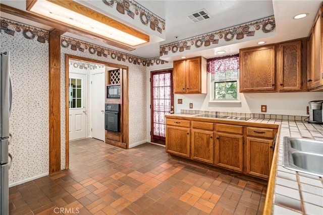 kitchen featuring tile countertops, visible vents, wallpapered walls, brick floor, and black appliances