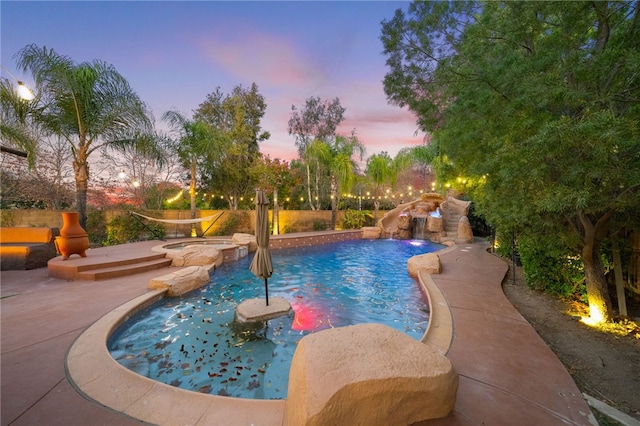 pool at dusk with a patio, a pool with connected hot tub, a water slide, and fence