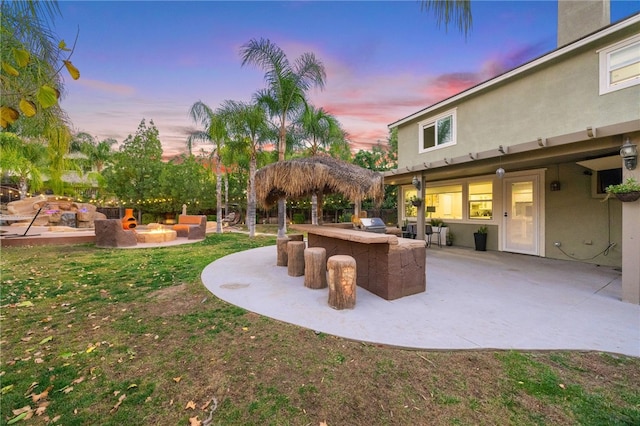 patio terrace at dusk featuring a lawn, a fire pit, and exterior kitchen