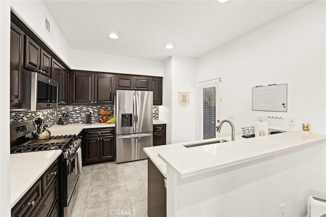 kitchen featuring dark brown cabinets, decorative backsplash, a peninsula, stainless steel appliances, and a sink