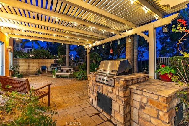 patio at night featuring area for grilling, fence, a pergola, and an outdoor kitchen