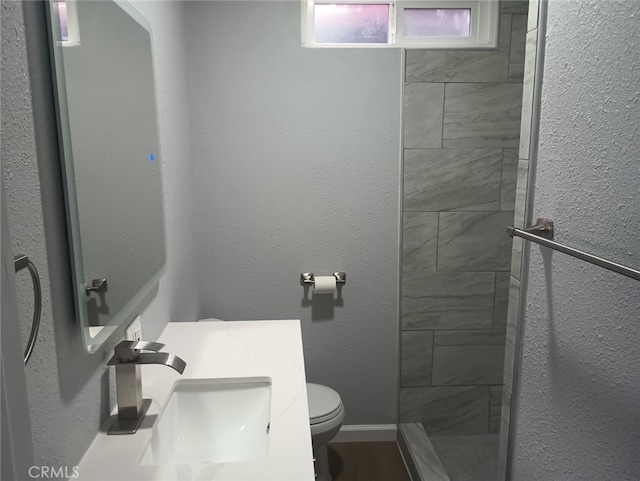 full bath with baseboards, tiled shower, a sink, toilet, and a textured wall