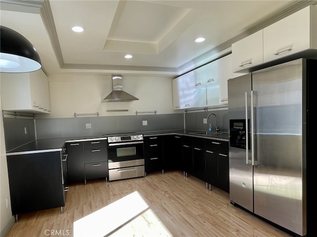 kitchen featuring a sink, a tray ceiling, stainless steel appliances, wall chimney range hood, and dark cabinets