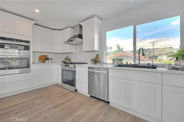 kitchen with appliances with stainless steel finishes, light wood-type flooring, wall chimney exhaust hood, and light countertops