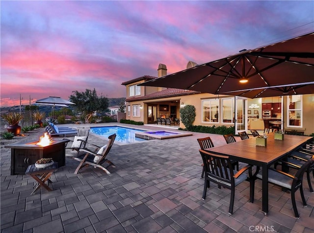 pool at dusk featuring a pool with connected hot tub, a fire pit, outdoor dining area, and a patio