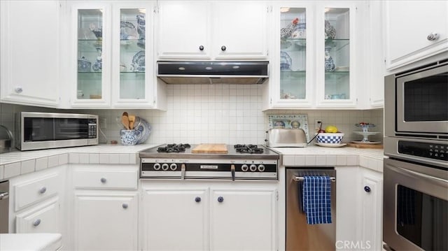 kitchen featuring under cabinet range hood, backsplash, tile countertops, stainless steel appliances, and white cabinets