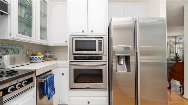 kitchen with tasteful backsplash, white cabinetry, stainless steel appliances, glass insert cabinets, and tile counters