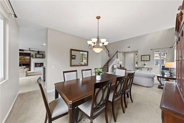 dining space featuring stairway, baseboards, a fireplace, ceiling fan with notable chandelier, and light colored carpet