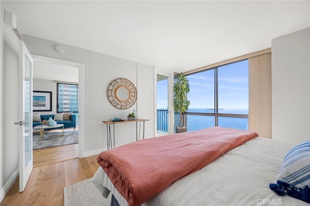 bedroom featuring light wood-type flooring, a water view, floor to ceiling windows, baseboards, and access to exterior