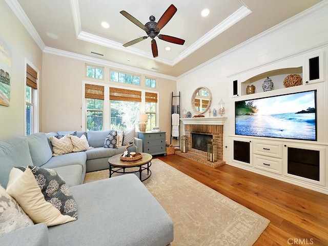 living room with a brick fireplace, ornamental molding, wood finished floors, a raised ceiling, and a ceiling fan