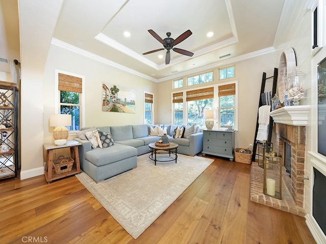 living area with a ceiling fan, light wood finished floors, crown molding, a raised ceiling, and a brick fireplace