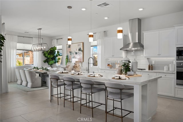 kitchen with visible vents, a center island with sink, backsplash, white cabinets, and wall chimney range hood