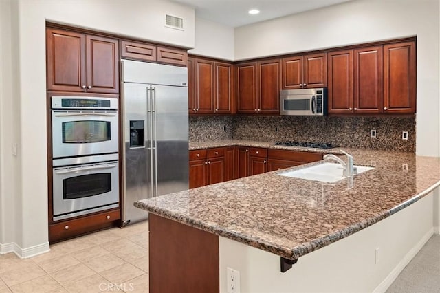 kitchen with a sink, backsplash, stainless steel appliances, dark stone counters, and a peninsula