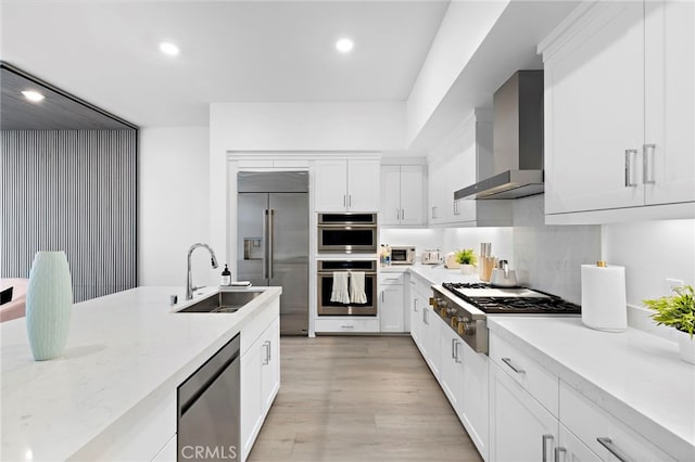 kitchen featuring light wood finished floors, a sink, stainless steel appliances, white cabinetry, and wall chimney exhaust hood