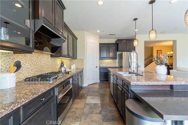 kitchen with a center island with sink, a sink, under cabinet range hood, tasteful backsplash, and appliances with stainless steel finishes