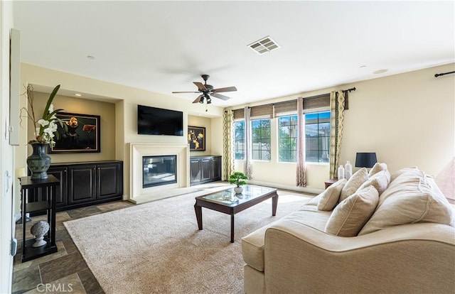 living room with ceiling fan, visible vents, a glass covered fireplace, and stone finish flooring