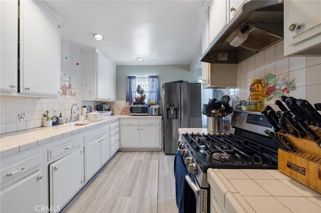 kitchen featuring backsplash, under cabinet range hood, tile countertops, white cabinets, and stainless steel appliances