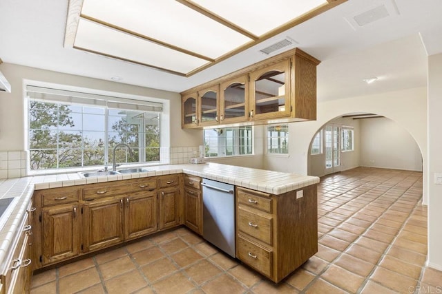 kitchen featuring brown cabinetry, dishwasher, a peninsula, arched walkways, and a sink