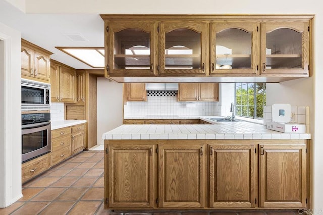 kitchen with a sink, tasteful backsplash, appliances with stainless steel finishes, a peninsula, and glass insert cabinets