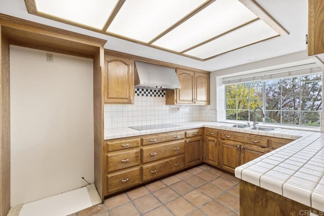 kitchen with brown cabinets, black electric cooktop, a sink, wall chimney exhaust hood, and decorative backsplash