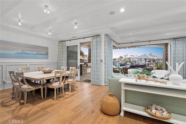 dining area featuring beam ceiling, a decorative wall, light wood-style flooring, and wainscoting