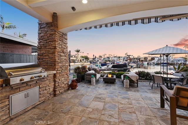 patio terrace at dusk with area for grilling, an outdoor kitchen, outdoor dining space, and a water view