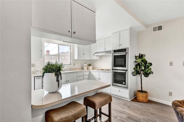 kitchen with a breakfast bar area, visible vents, light wood-style flooring, under cabinet range hood, and double oven