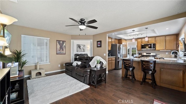 living area featuring a ceiling fan, dark wood-style floors, baseboards, a tray ceiling, and a textured ceiling