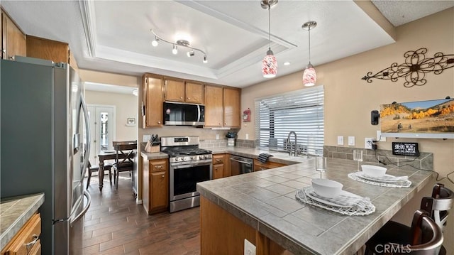 kitchen featuring a peninsula, stainless steel appliances, pendant lighting, a raised ceiling, and brown cabinets