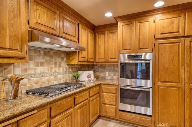 kitchen featuring brown cabinets, under cabinet range hood, stainless steel appliances, decorative backsplash, and light stone countertops