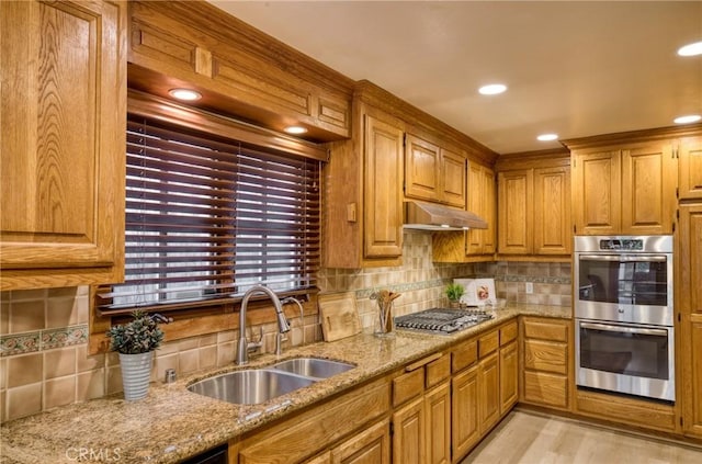 kitchen featuring a sink, light stone counters, under cabinet range hood, stainless steel appliances, and brown cabinetry