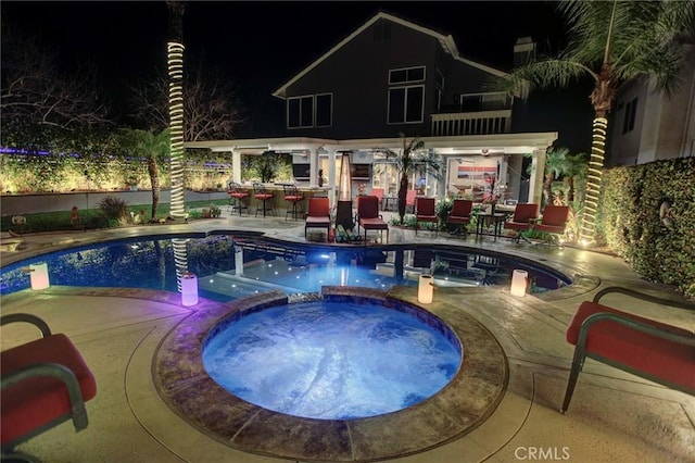 pool at night featuring a patio area, a pool with connected hot tub, and outdoor dry bar