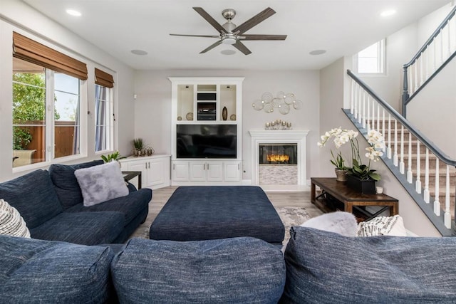 living area featuring light wood finished floors, stairway, recessed lighting, a glass covered fireplace, and a ceiling fan