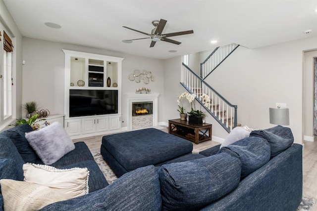 living room with a glass covered fireplace, light wood-style floors, baseboards, ceiling fan, and stairs