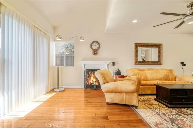 sitting room with baseboards, vaulted ceiling, a warm lit fireplace, wood finished floors, and a ceiling fan