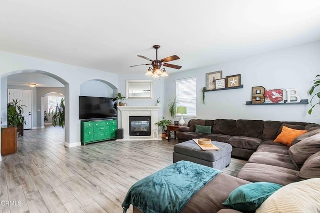 living area featuring light wood-type flooring, a glass covered fireplace, arched walkways, baseboards, and ceiling fan