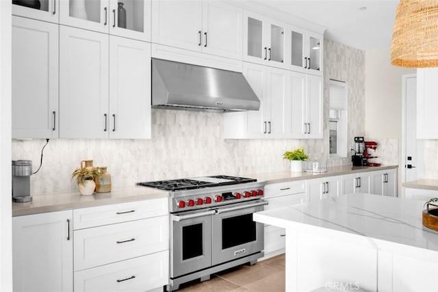 kitchen with light stone counters, range with two ovens, decorative backsplash, white cabinetry, and wall chimney exhaust hood