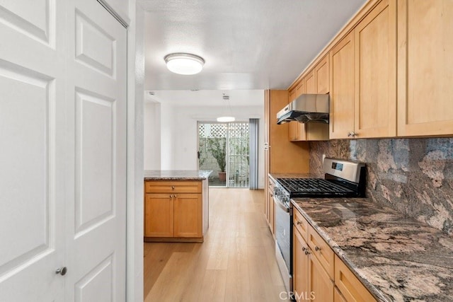 kitchen with light wood-style flooring, under cabinet range hood, backsplash, dark stone counters, and stainless steel range with gas cooktop