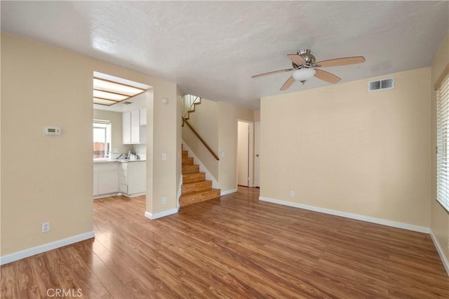 unfurnished living room with a ceiling fan, visible vents, light wood finished floors, baseboards, and stairs