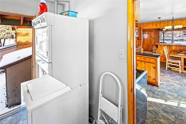 laundry area with wooden walls, laundry area, stone finish floor, stacked washer / dryer, and a notable chandelier