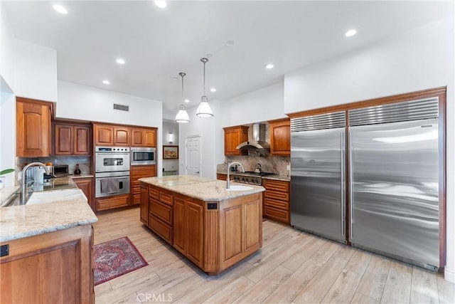 kitchen featuring light wood-style flooring, a kitchen island with sink, a sink, wall chimney range hood, and built in appliances