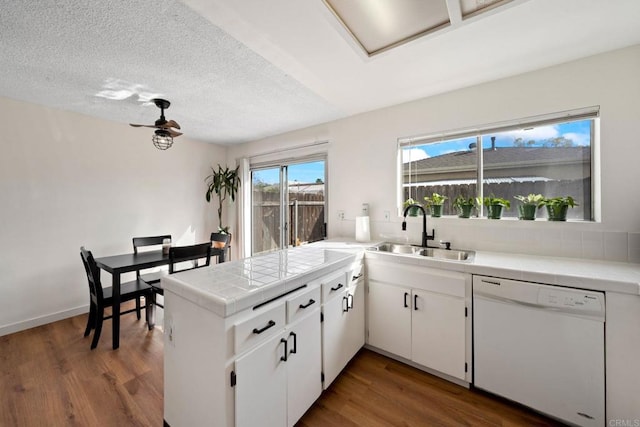 kitchen featuring tile counters, dishwasher, a peninsula, wood finished floors, and a sink