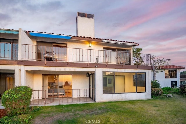 back of property at dusk with stucco siding, a lawn, a balcony, and a chimney