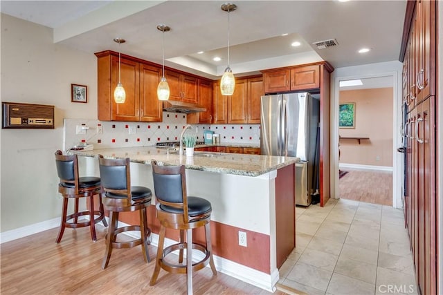 kitchen with light stone counters, under cabinet range hood, backsplash, freestanding refrigerator, and a peninsula